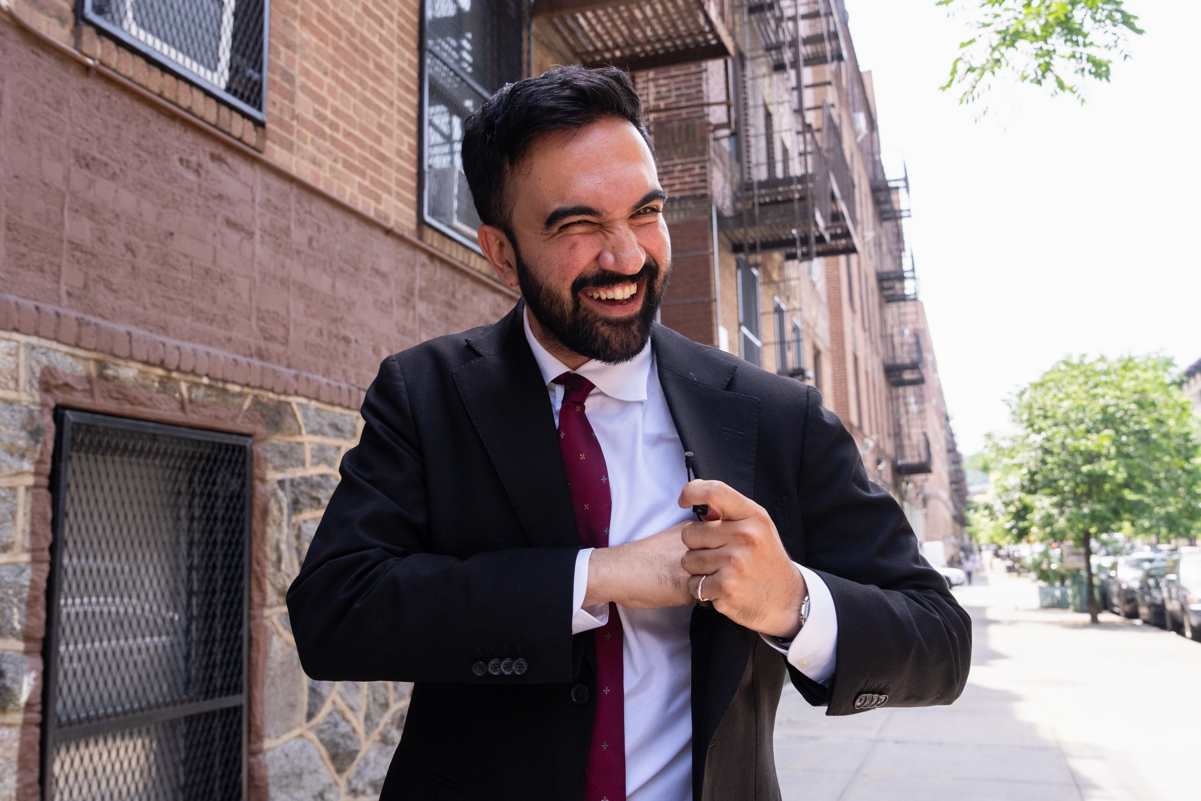 Man in suit and tie laughs while adjusting jacket on a city sidewalk