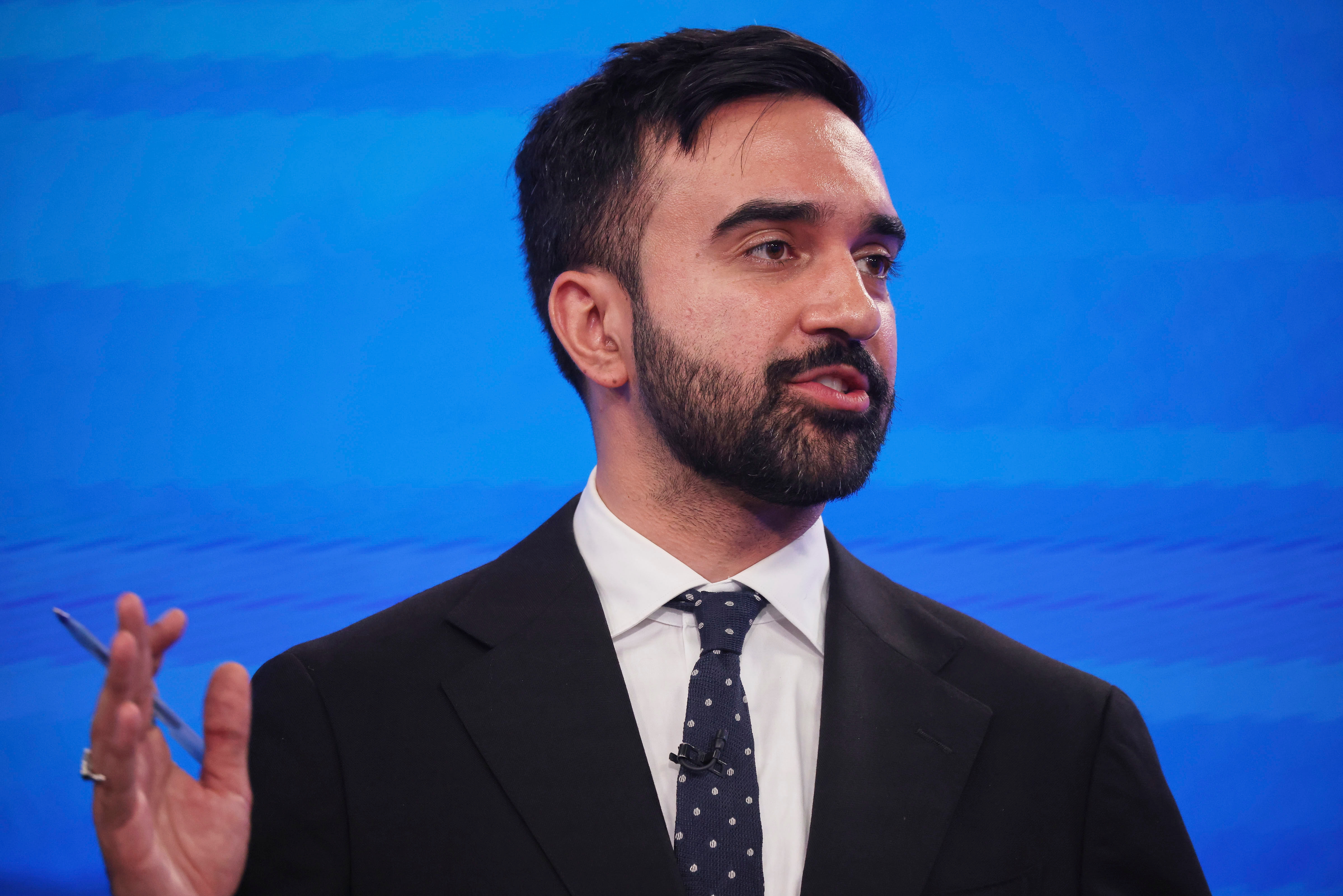 Person wearing a suit with a polka-dot tie, gesturing while speaking on stage or in front of an audience