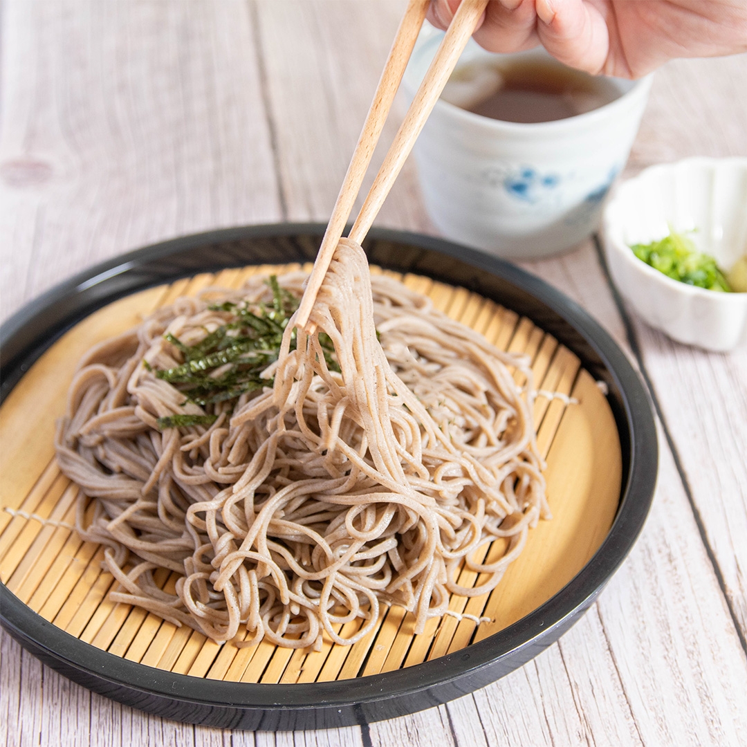 手がそばを箸で持ち上げています。竹のざるには盛られたそばがあり、奥にはつゆと刻み葱が見えます。