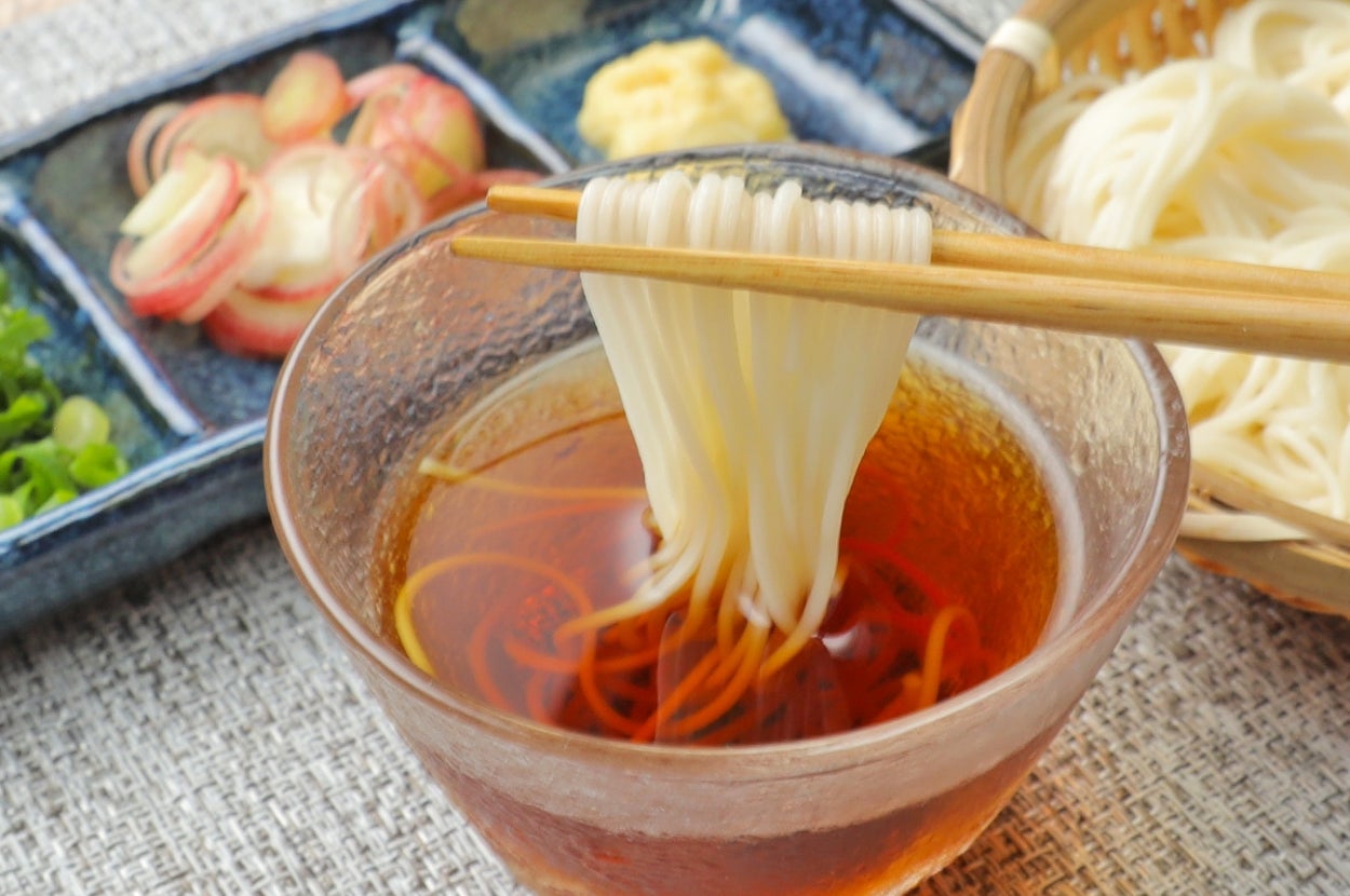そうめんと薬味の盛り付け。ガラスの器にめんつゆ、箸でそうめんを持ち上げているシーン。薬味は別皿に盛られています。