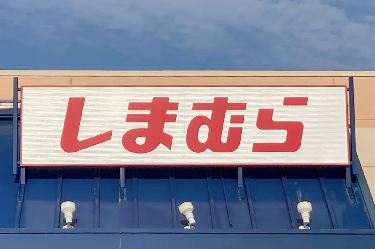 A store sign with Japanese text reading "しまむら" on a building facade. Blue awning below the sign