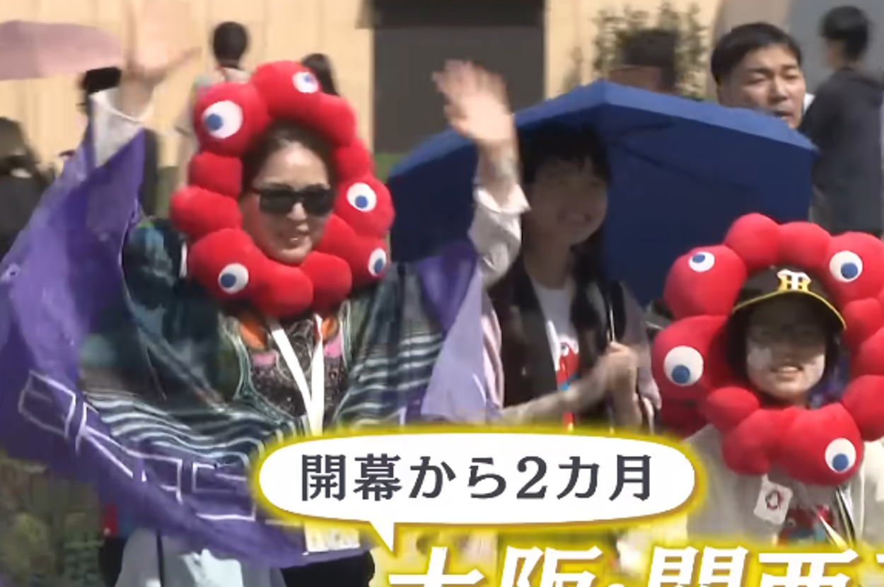 グループが大阪・関西万博でユニークな衣装を着てカメラに向かって手を振っている。