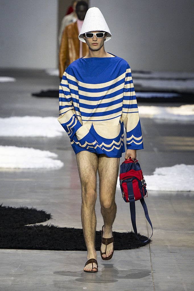Model on runway wears oversized striped top, white hat, sunglasses, holding red bag, and slippers. Fashion show setting with textured floor elements