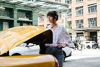 Person standing beside an open car trunk, wearing a casual blouse and jeans, in an urban street setting with buildings and people in the background