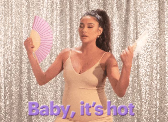 A person wearing a sleeveless dress uses two fans to cool down, with text reading &quot;Baby, it&#x27;s hot&quot; in front of a sparkly backdrop