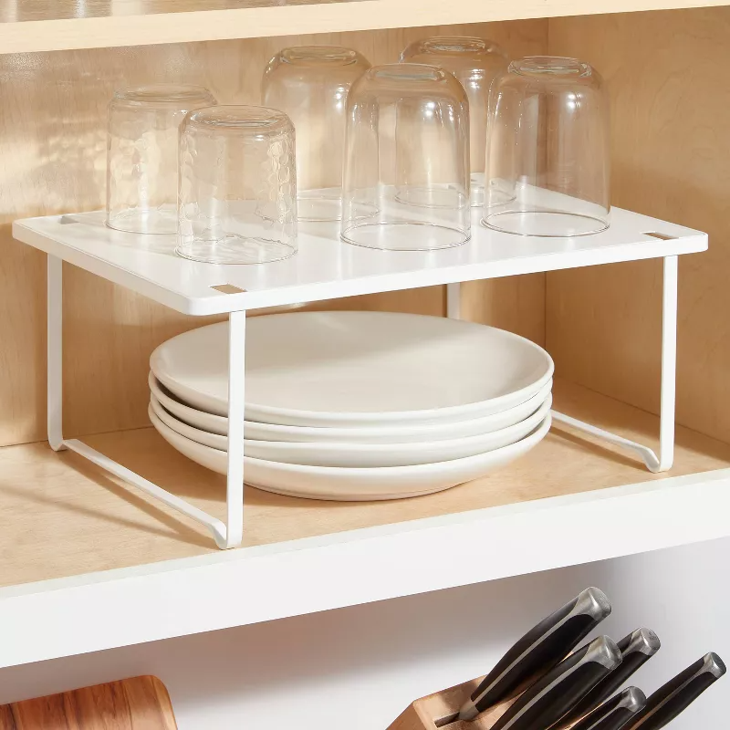 Cabinet shelf organizer with glasses on top and plates below, maximizing storage space. Knives are visible in a block nearby