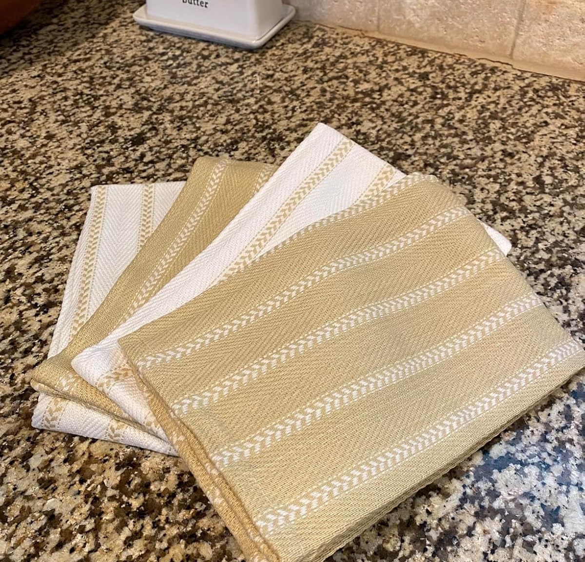 Reviewer's photo of four folded kitchen towels with simple striped patterns on a speckled granite countertop. A butter dish is in the background