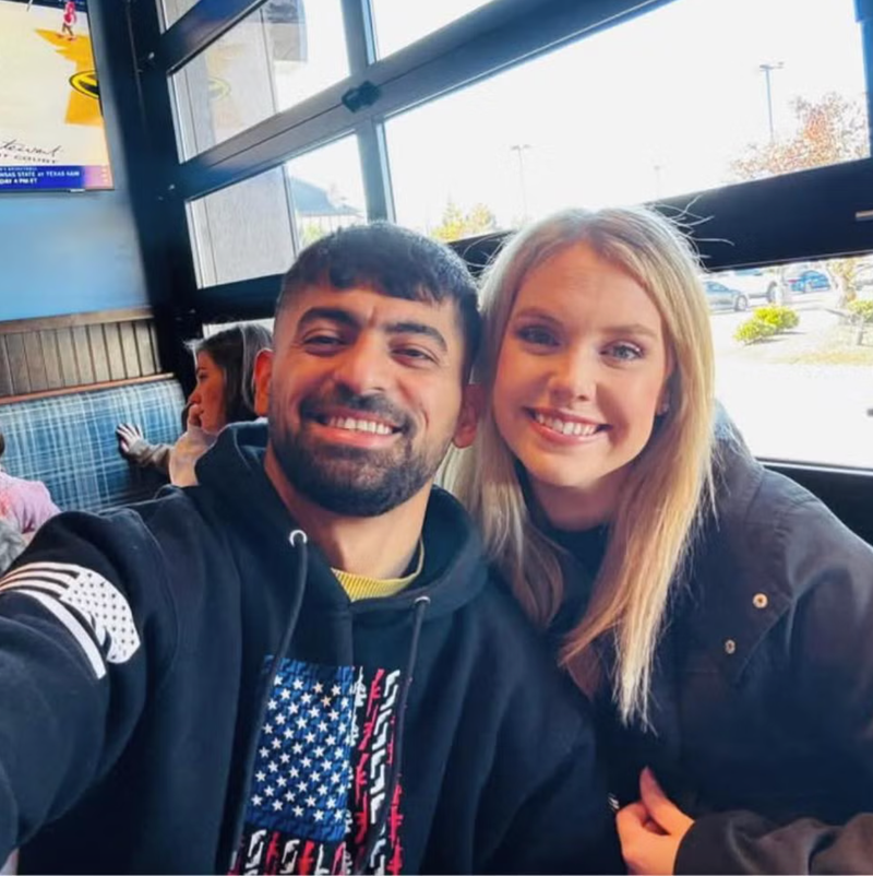 Man and woman smiling, sitting in a casual indoor setting. Man wears a hoodie with an American flag design; woman wears a dark jacket