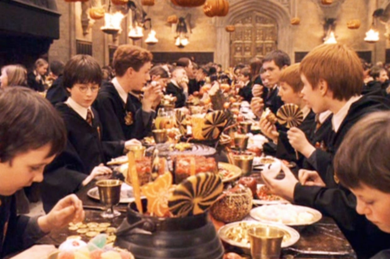 Hogwarts students dressed in school uniforms enjoy a festive meal at a long dining table in the Great Hall with floating pumpkins above