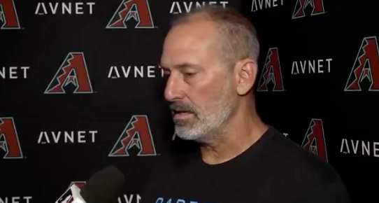 A man being interviewed at a sports event, wearing a casual t-shirt, with a logo backdrop behind him