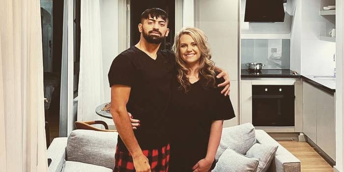 A man and a woman stand in a modern kitchen, both wearing casual black outfits. The man has his arm around the woman, who is smiling at the camera