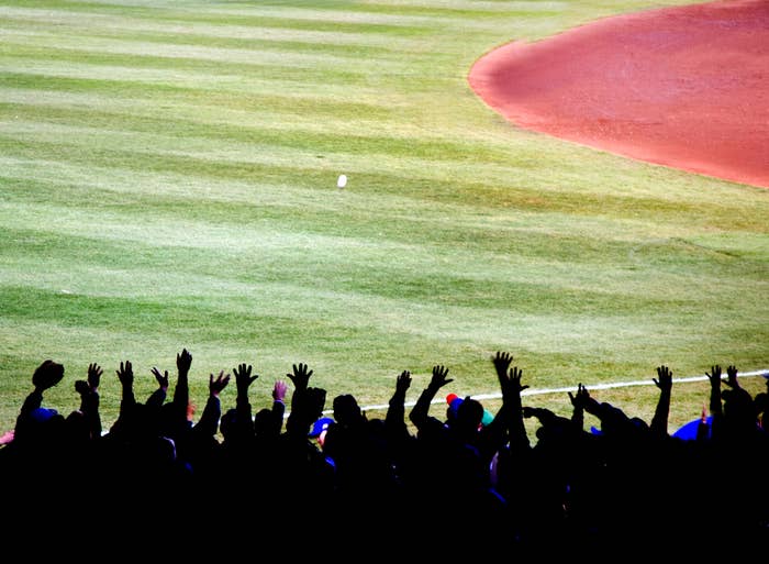 Fans cheer at a baseball field, with raised hands silhouetted against the outfield grass and infield dirt