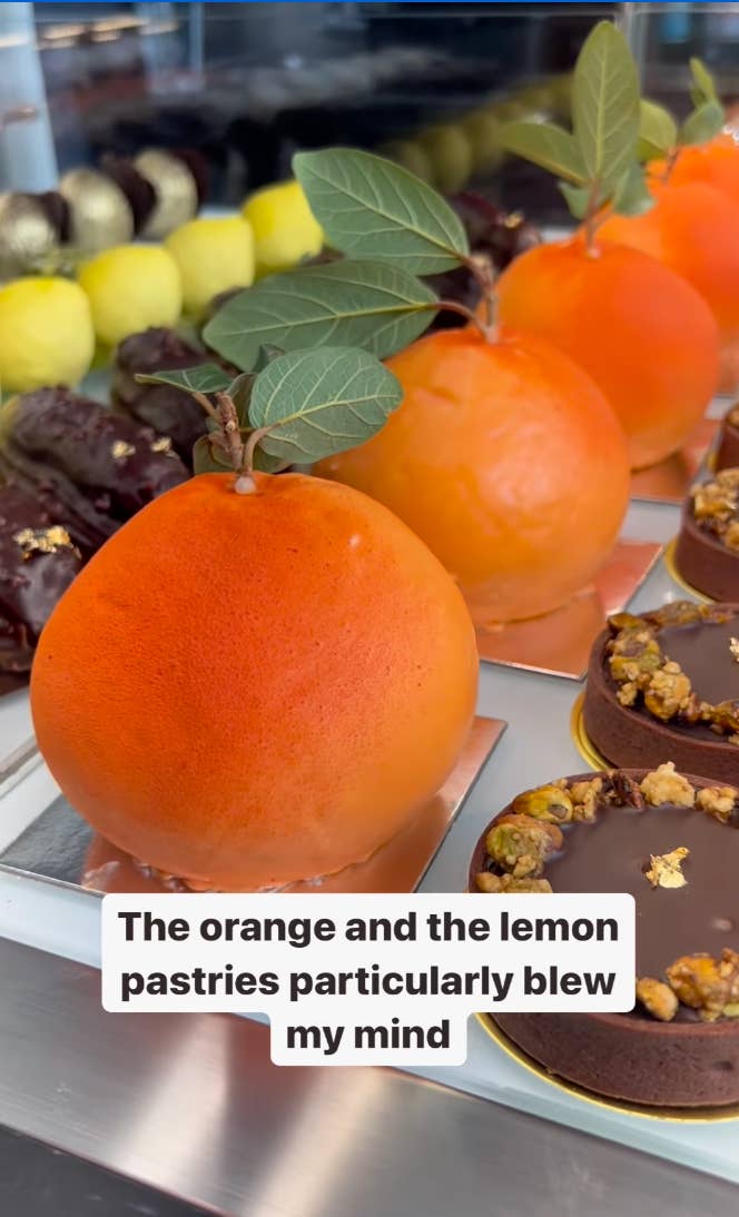 Close-up of vibrant cakes shaped like oranges with leaves, alongside various pastries. A text overlay reads, &quot;The orange and the lemon pastries particularly blew my mind.&quot;