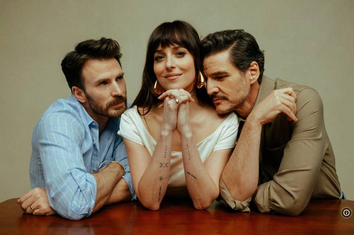 Three people pose with relaxed expressions, leaning on a table. The person in the center wears a white top and rests hands under their chin