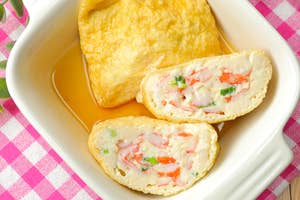 白い皿に入った焼き豆腐と野菜が詰まった揚げ物がピンクのチェック柄の布の上に置かれている。端に箸がある。