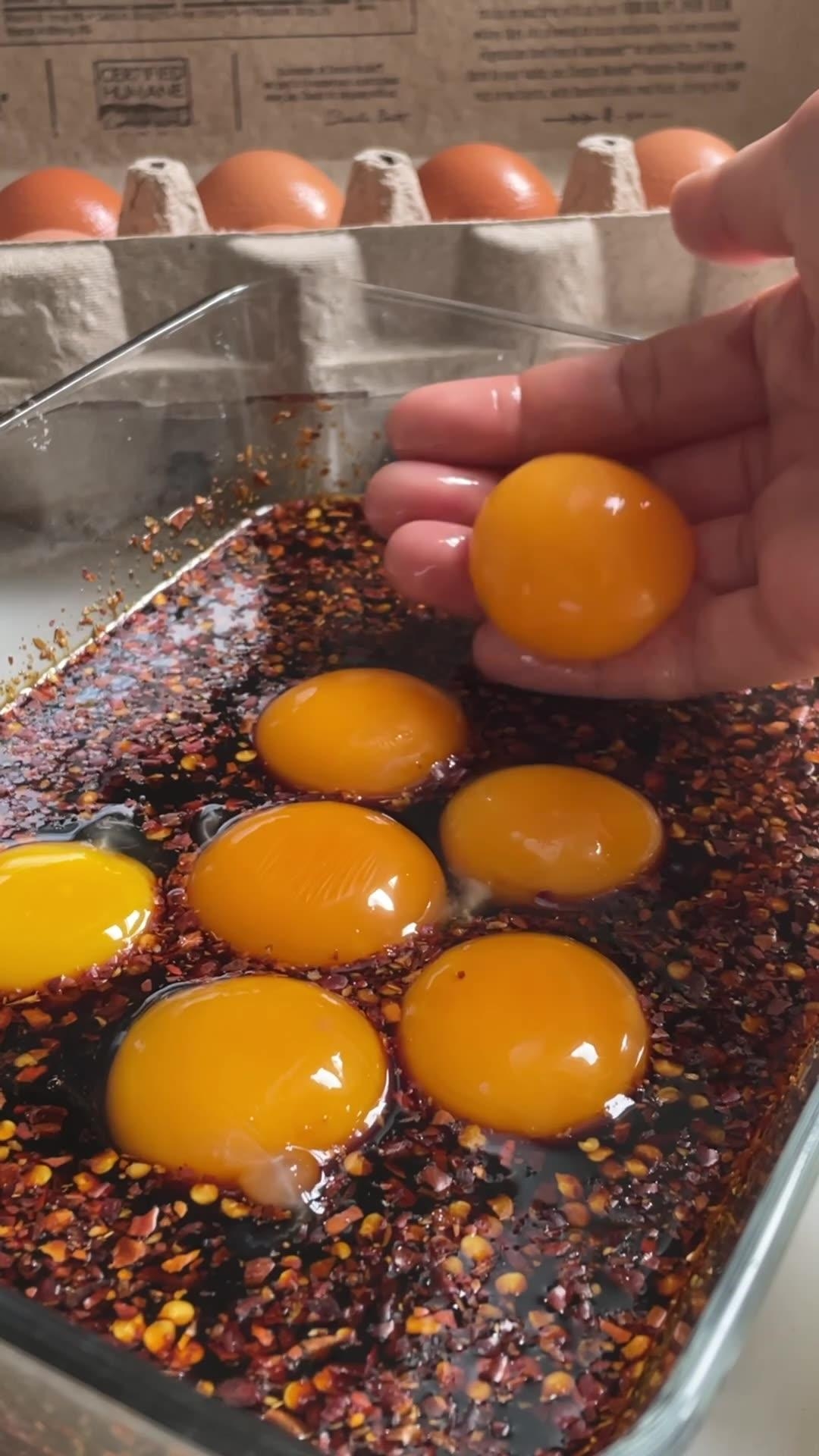 Six egg yolks in a glass dish with soy sauce and chili flakes. A hand holds one yolk above the mixture