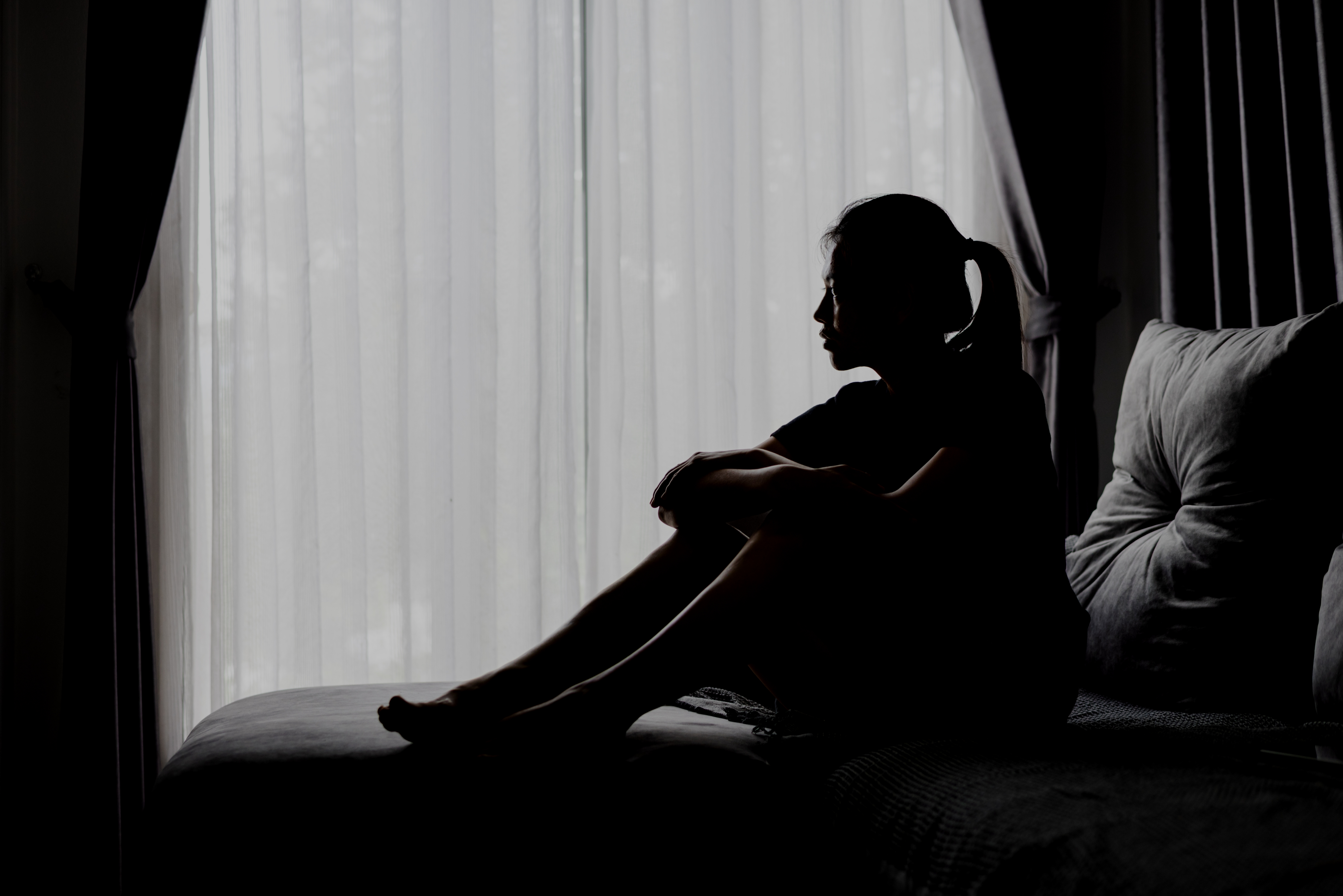 Person som sitter på en sofa, stirrer gjennomtenkt et vindu med rene gardiner, skaper en kontemplativ og rolig atmosfære