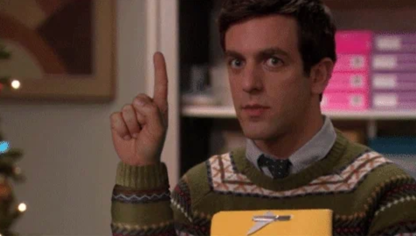 Personen holder et utklippstavle, gestikulerer med en pekefinger hevet, iført en mønstret genser og slips, i et kontorinnstilling