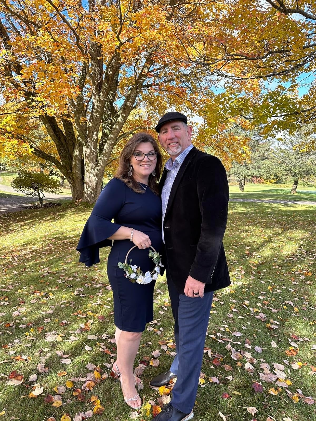 A couple stands smiling in a park with autumn leaves. The woman wears a stylish dress and holds a small floral bouquet; the man wears a blazer and cap