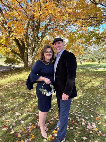 A couple stands smiling in a park with autumn leaves. The woman wears a stylish dress and holds a small floral bouquet; the man wears a blazer and cap