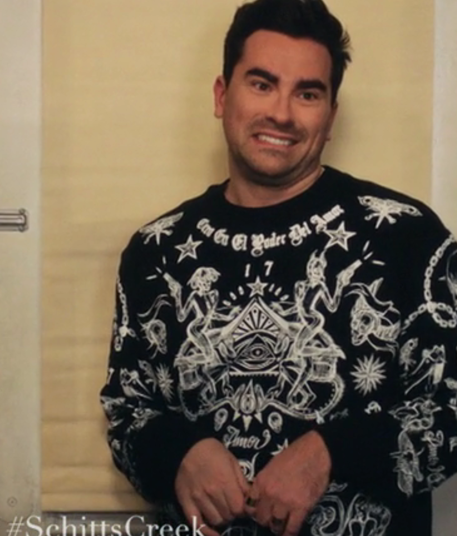 Person som står tafatt med et nervøst smil, iført en detaljert mønstret genser med intrikate design og symboler