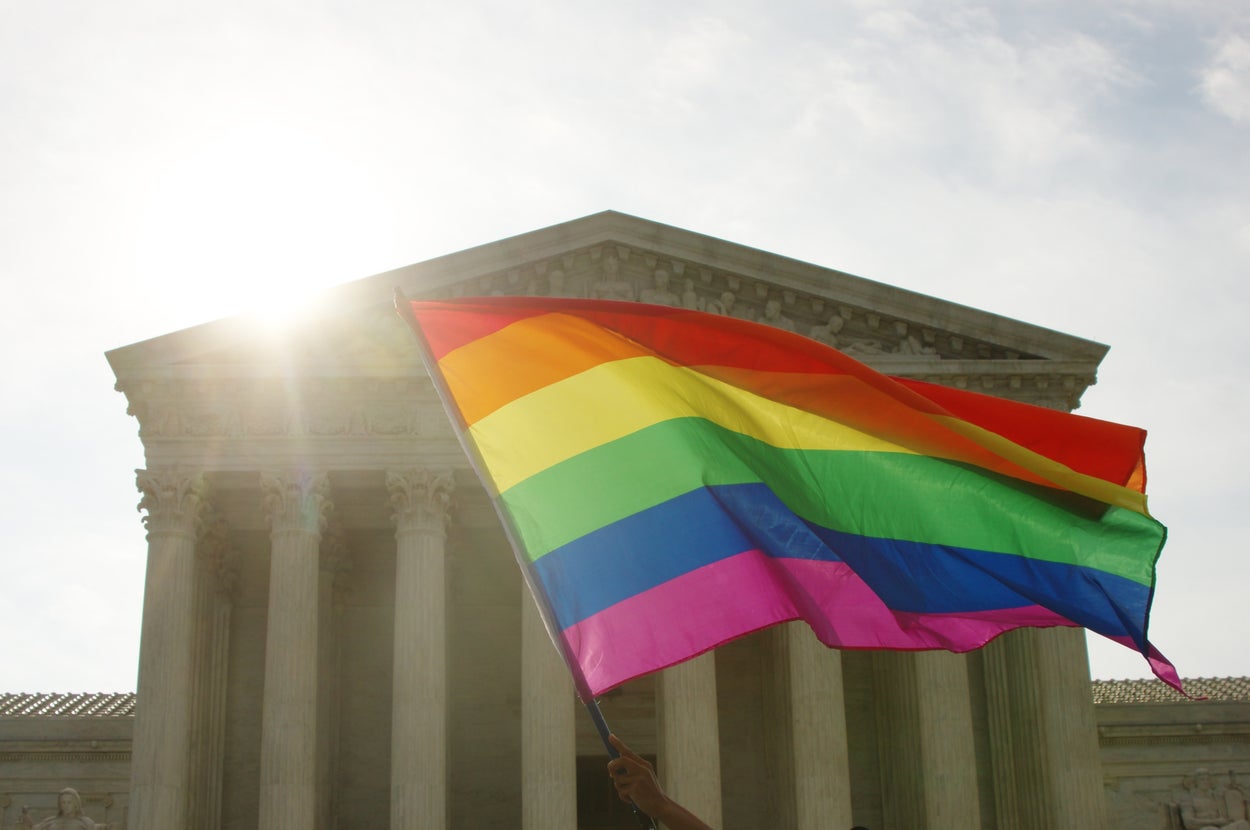 Person holding a rainbow flag in front of a classical building with columns, symbolizing LGBTQ+ pride and advocacy for equal rights