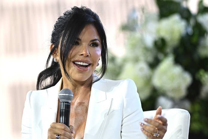 Lauren Sánchez with long hair in a stylish updo, wearing a blazer, speaking into a microphone and gesturing while seated against a blurred background