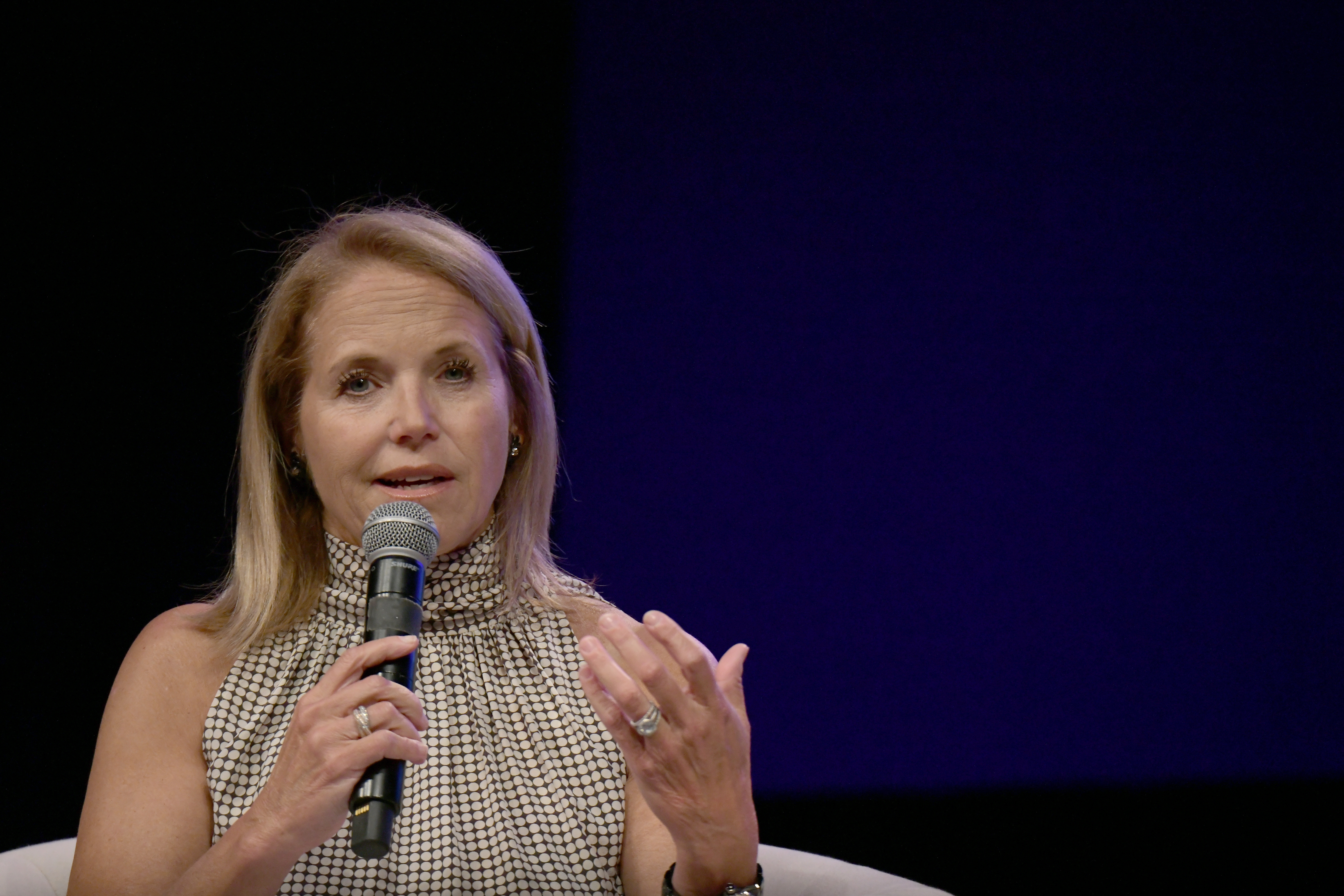 Katie Couric speaks into a microphone while seated on stage, wearing a checkered sleeveless top
