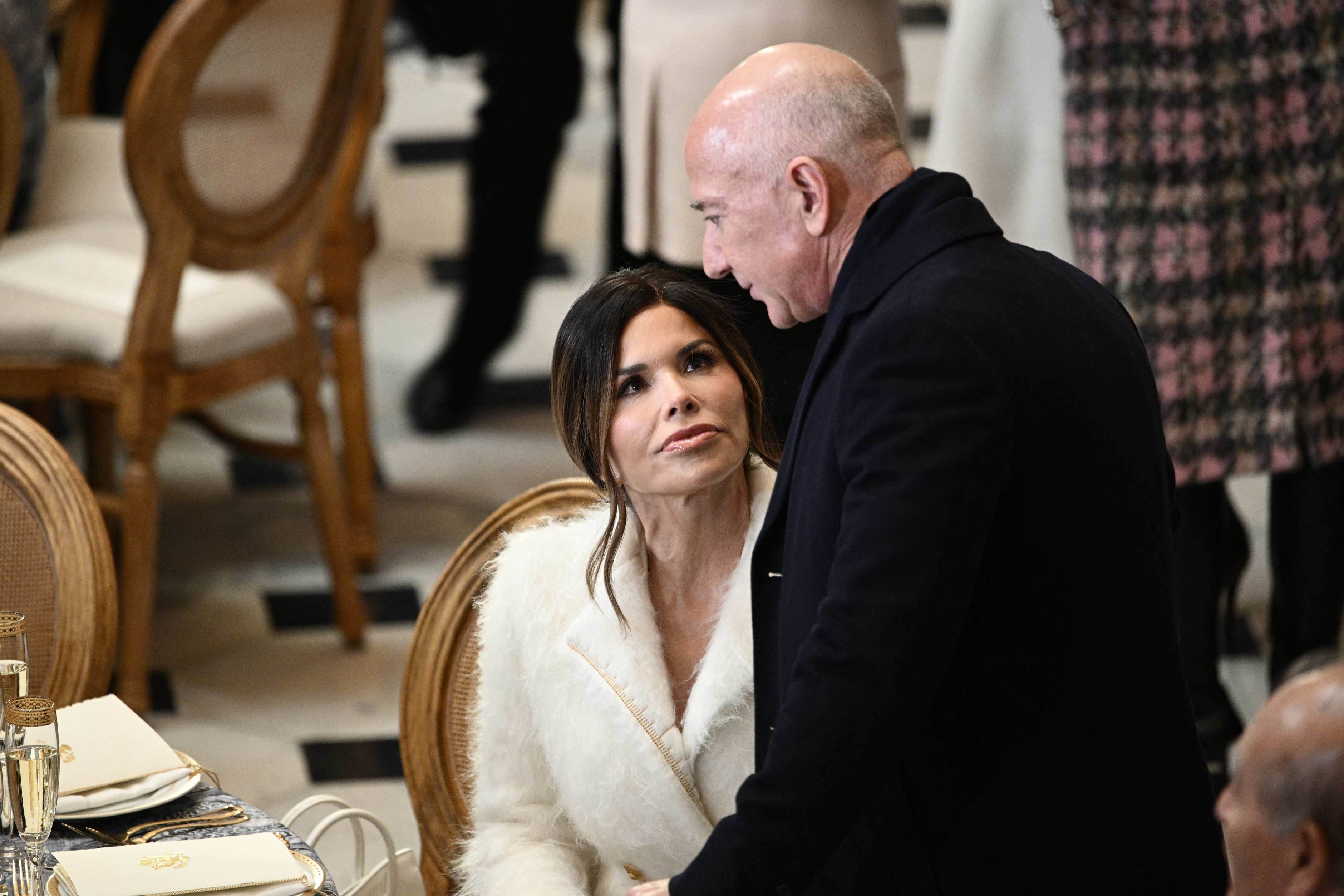 Lauren Sanchez sits at a dining table, looking up at Jeff Bezos standing beside her at a formal event