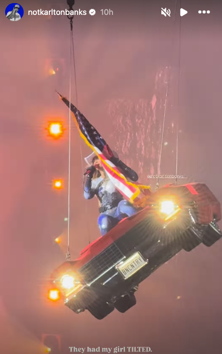 Performer in a shiny outfit is suspended from a car with an American flag
