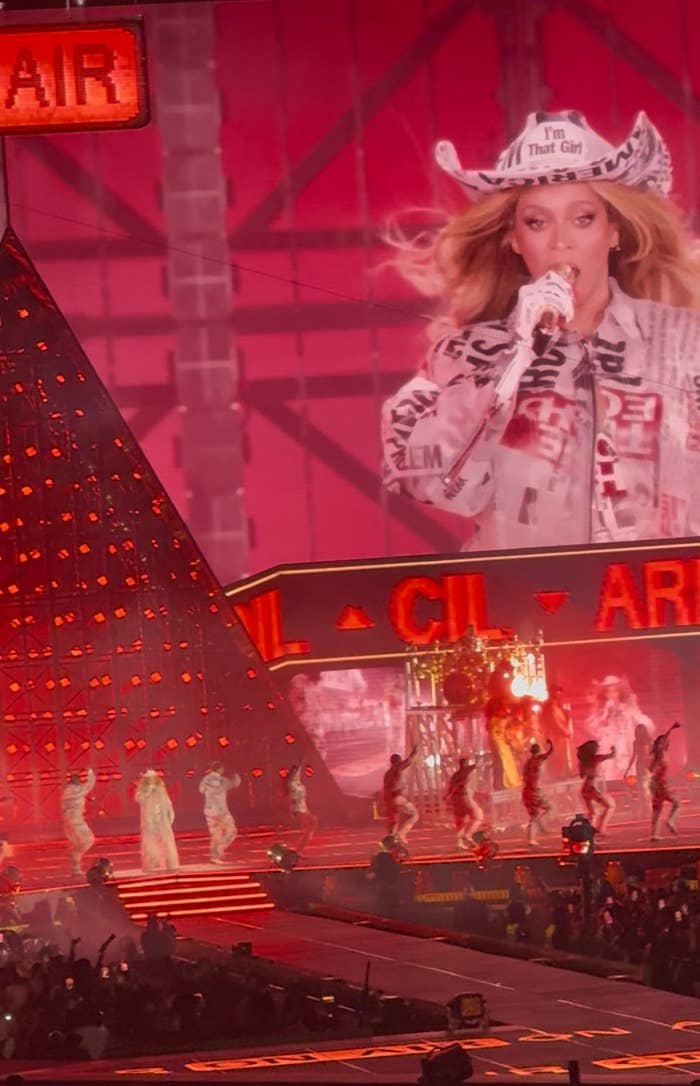 Performer on stage in a cowboy hat and patterned outfit, with dancers and vibrant stage lighting in the background