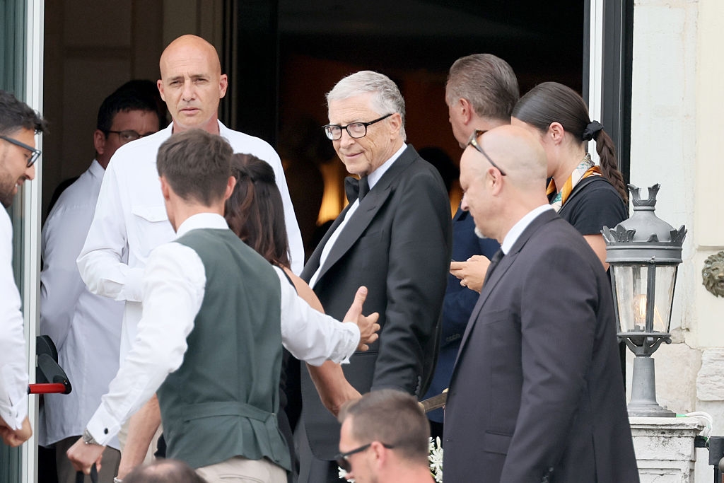 A group of people stand outside. A man in a tuxedo is prominently featured, surrounded by others in formal and casual attire