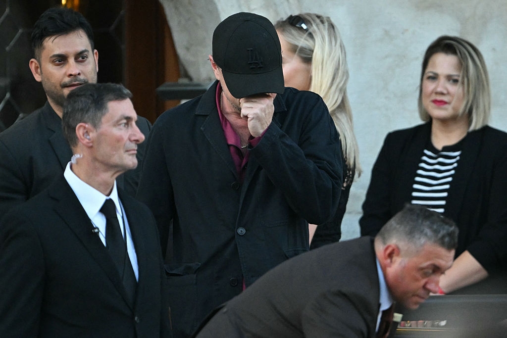 People at a formal event, one person covers their face. Others wear suits and dresses, conveying a somber mood