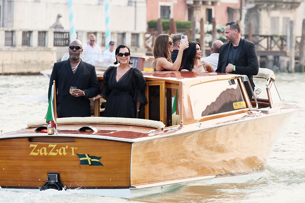 Group on a stylish wooden boat in a scenic canal setting, with some individuals posing and others taking photos