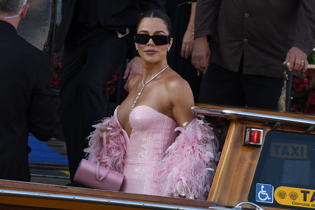 Person in glamorous, strapless dress with feathered shawl and necklace, exiting a boat. They wear sunglasses and carry a small handbag