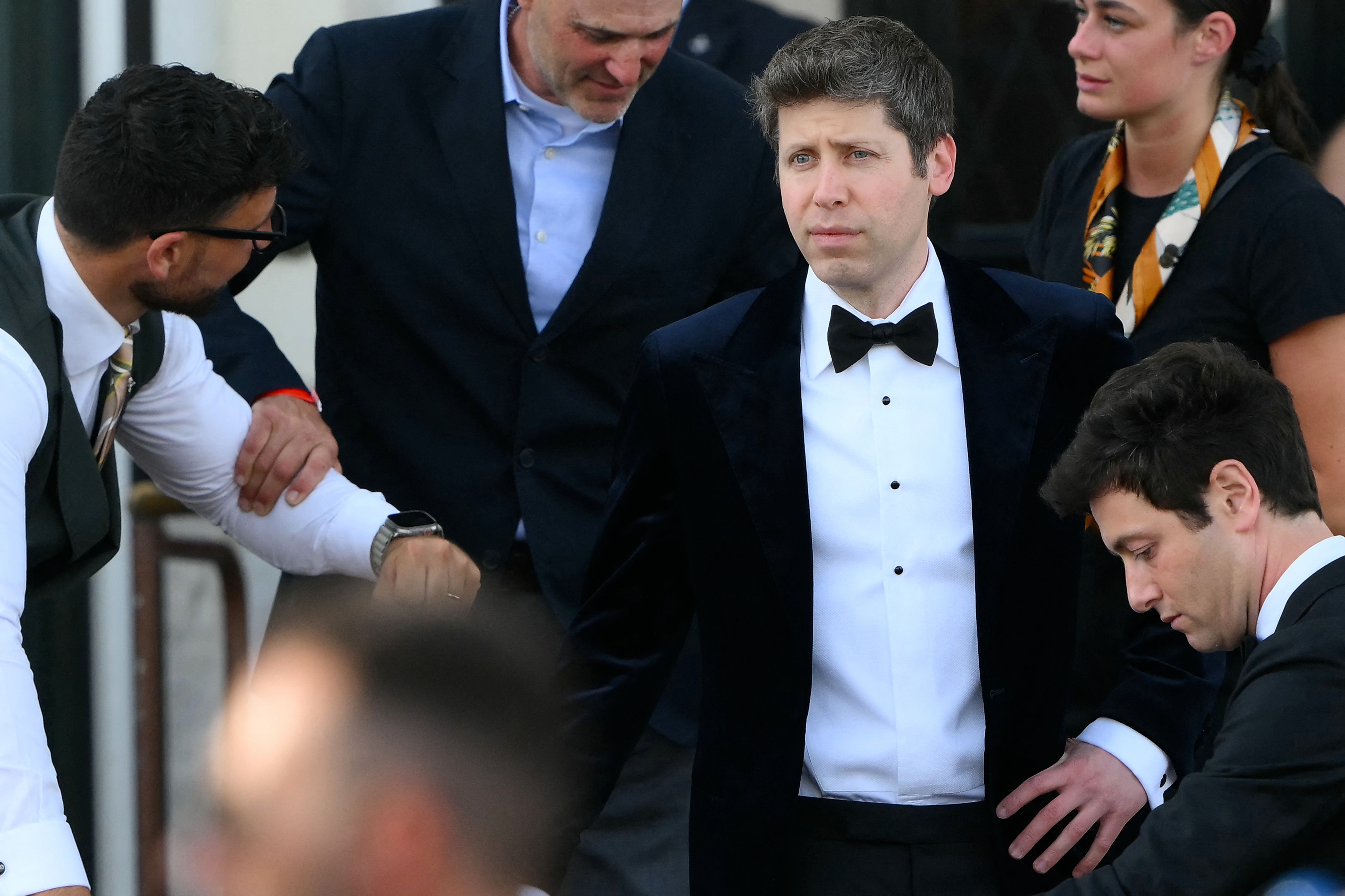 A man in a black tuxedo is surrounded by others at a formal event, likely on a red carpet