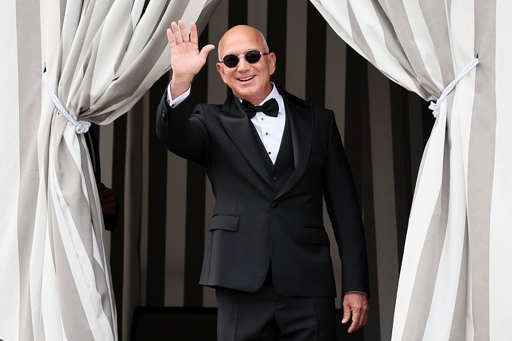 Man in a tailored tuxedo and sunglasses waves while standing between curtains
