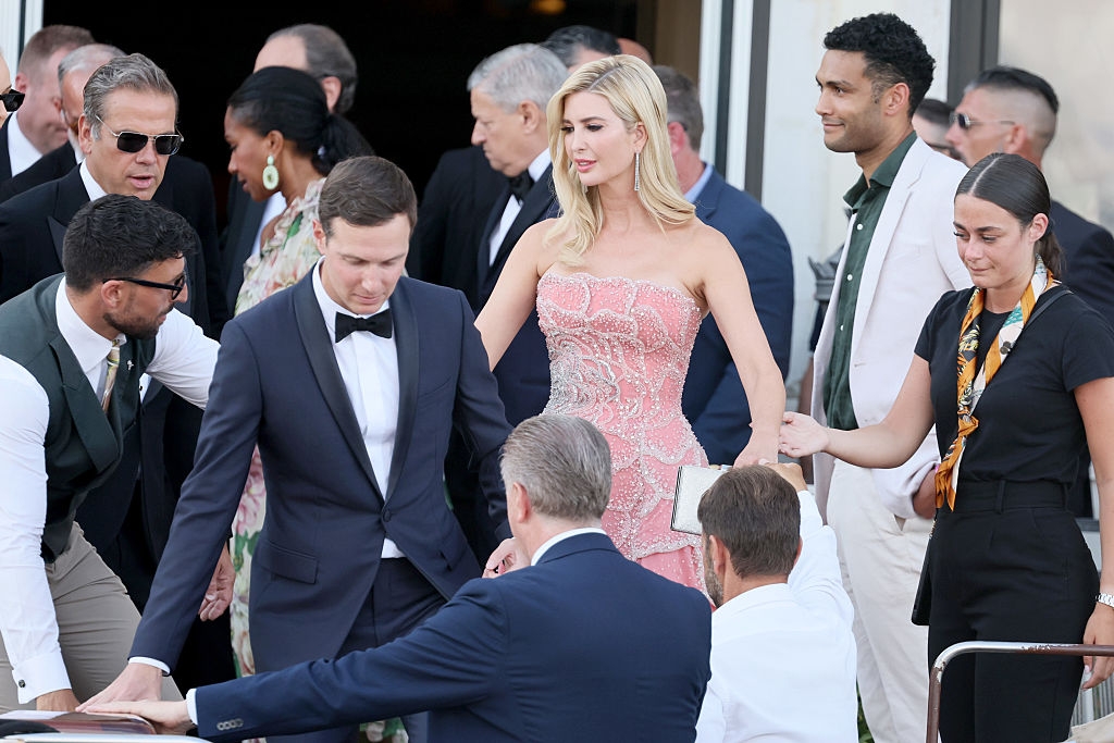 Group of people in formal attire at an event; woman in strapless, patterned gown and various men in suits. Interacting and socializing outdoors