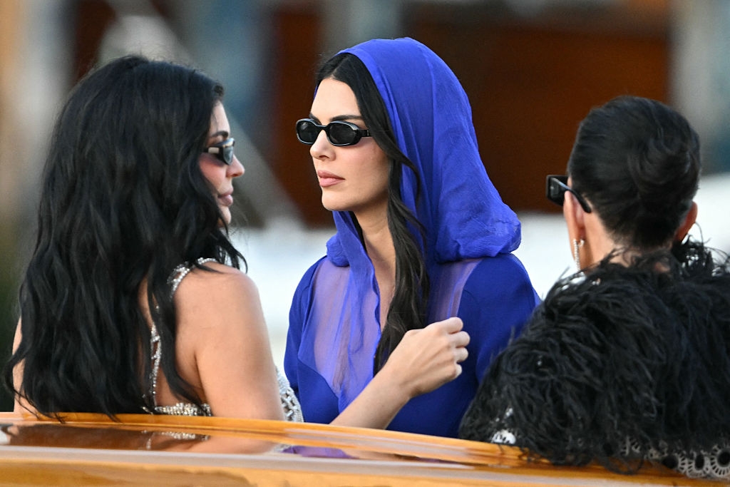 Three people are on a boat. The central person wears a sheer blue hooded outfit and black sunglasses, engaging with the others