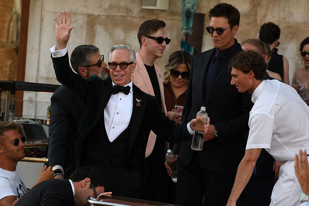 Group of people in formal wear and sunglasses interacting outdoors; one man waves enthusiastically