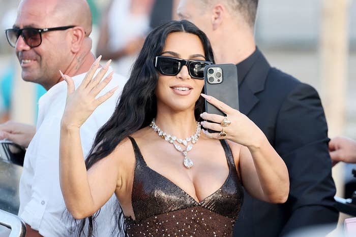 Person in a sparkling dress and sunglasses takes a selfie, showcasing their jewelry, surrounded by people at an outdoor event