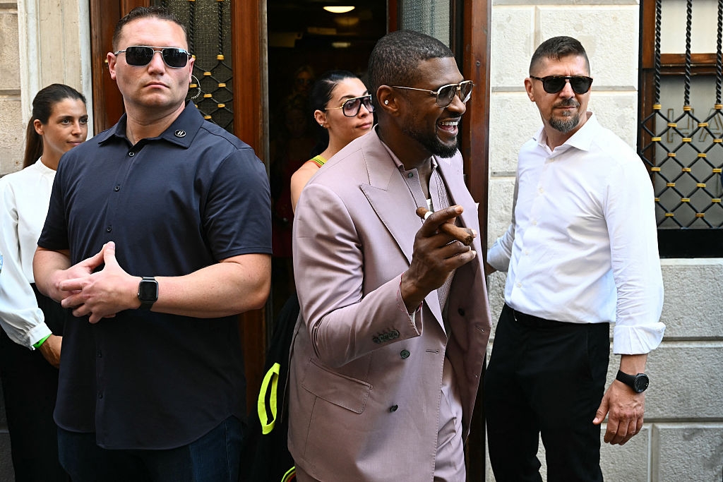 A well-dressed man in a fashionable suit smiles and gestures while surrounded by people, some appearing to be security personnel, outside a building