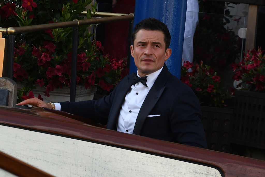A person in a formal tuxedo sits in a boat surrounded by flowers, appearing thoughtful