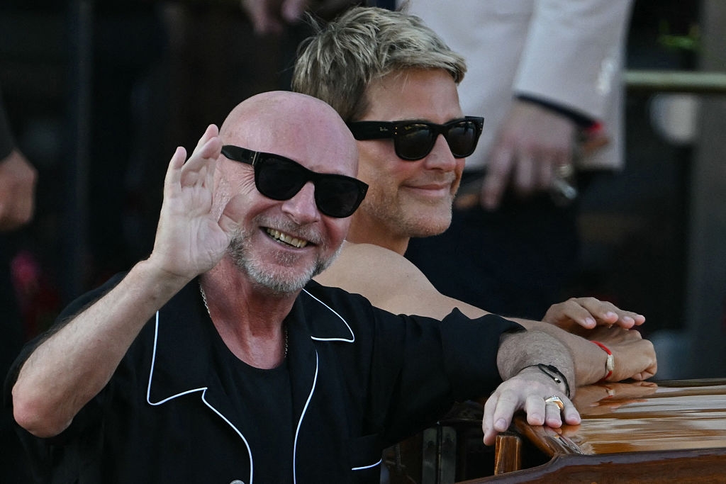 Two people wearing sunglasses, smiling and waving, sitting in a relaxed setting