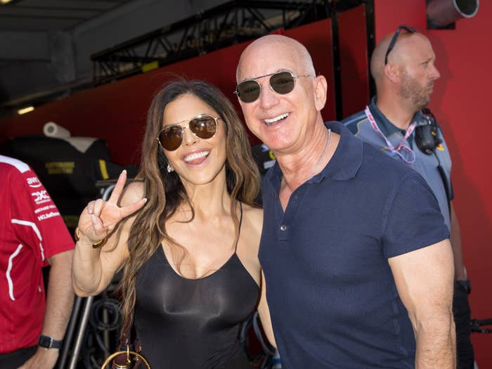 Two people wearing sunglasses smile and pose together; one flashes a peace sign. Casual attire, indoors with a red background and a standing bystander