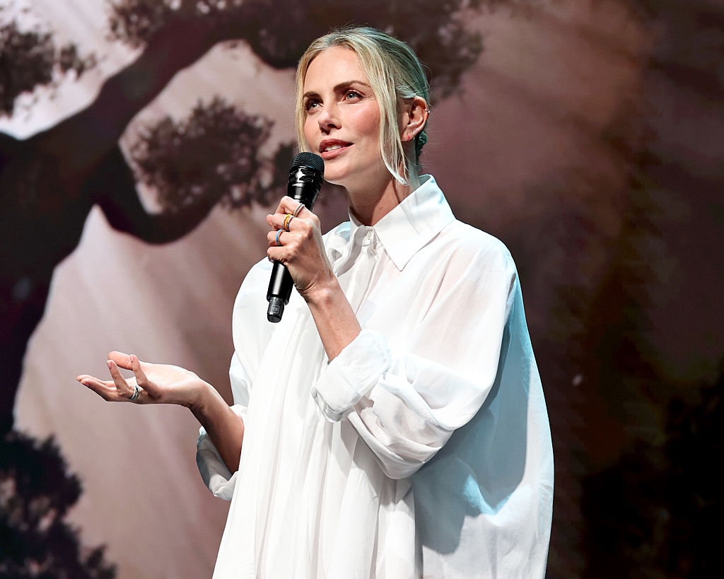Person in a flowing blouse speaks into a microphone on stage with a nature-themed background