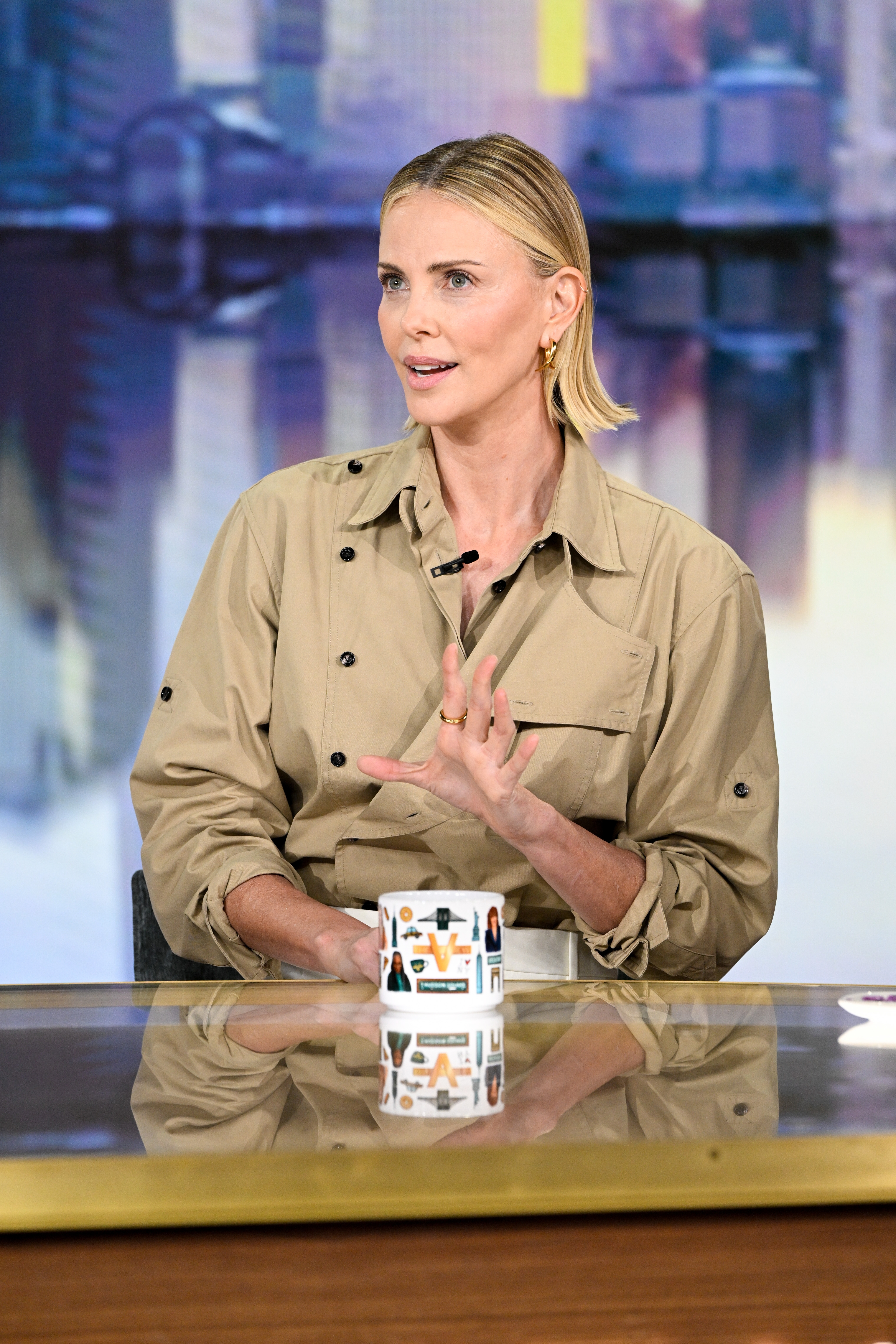 Person sitting at a table on a talk show, wearing a tan button-up shirt, speaking while gesturing with their hand