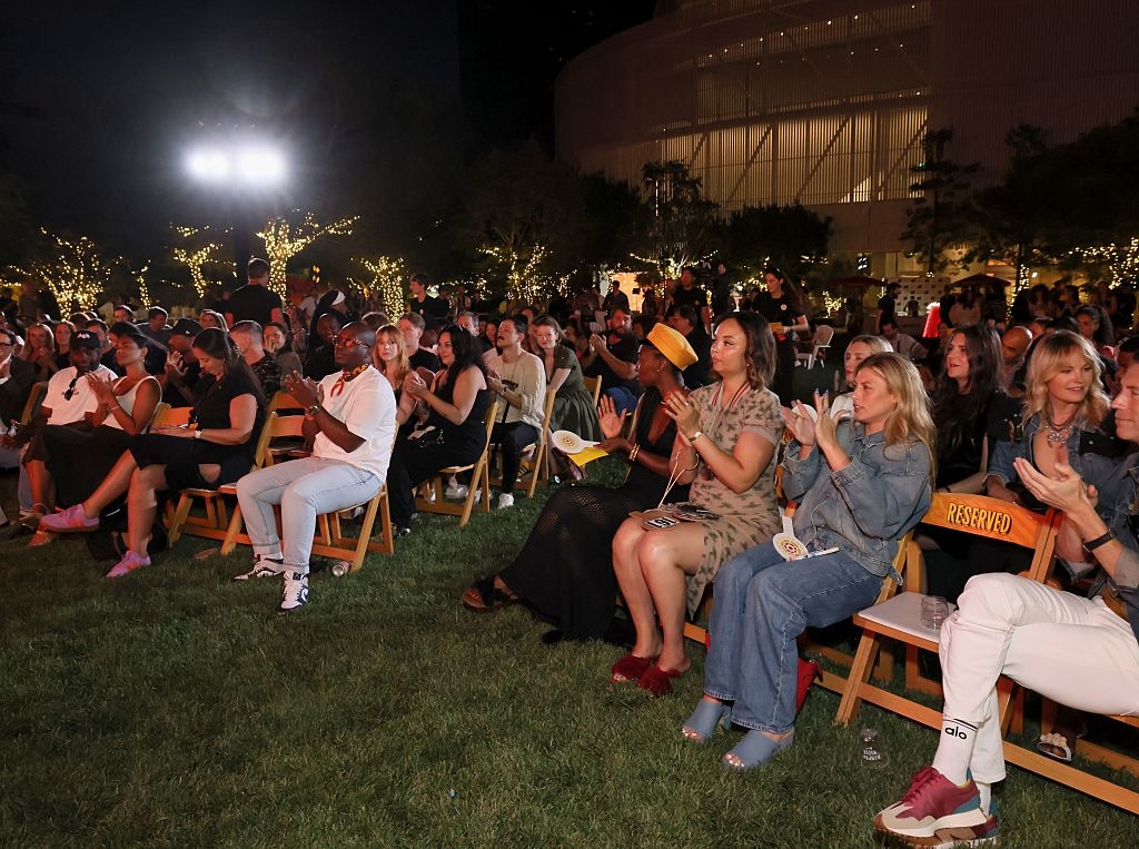 Audience seated outdoors, clapping at an evening event, some wearing casual attire, on folding chairs in a park-like setting with string lights