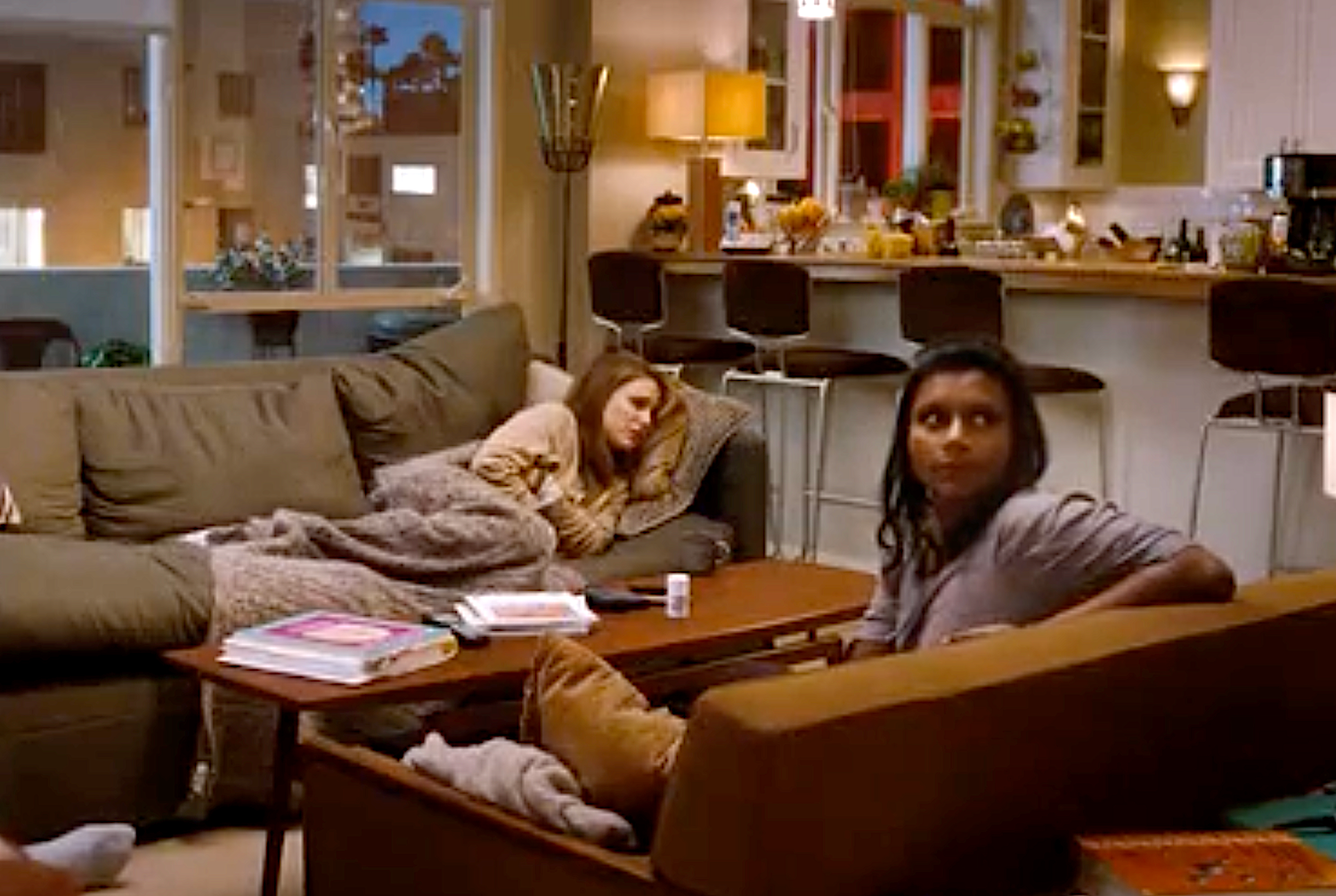 To personer slapper av i en koselig stue; Den ene ligger på en sofa med et teppe, mens den andre sitter i nærheten, og antyder en avslappet atmosfære