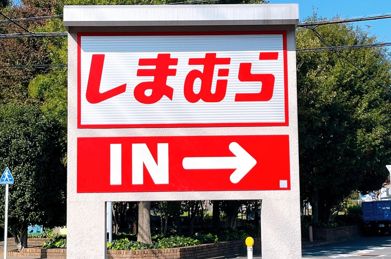 「しまむら IN→」と書かれた看板が表示されています。後ろに木々が見えます。